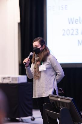 Image includes a light-skinned woman with long brown hair wearing a face mask speaking into a microphone.