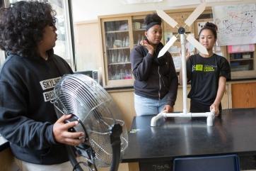 Three adolescents in a lab. One is holding a fan in front of two holding a propeller.