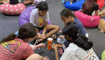 Four adolescent girls sitting in a circle pulling string against a cup.