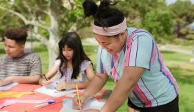 Three adolescents drawing on a table outdoors.